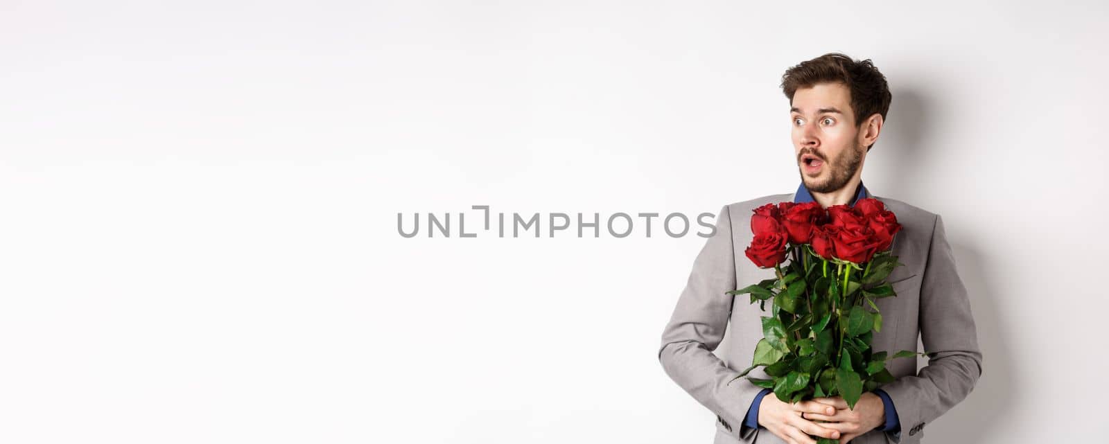 Handsome young man in suit holding red roses, looking left with surprised and startled expression, standing on Valentines day over white background by Benzoix