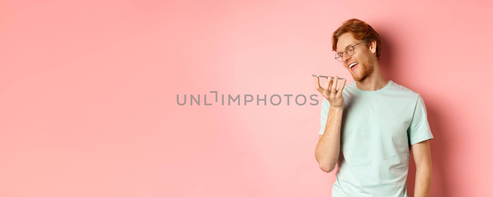 Happy young man with red hair, smiling pleased while record voice message on smartphone, talking to virtual assistant, standing over pink background.