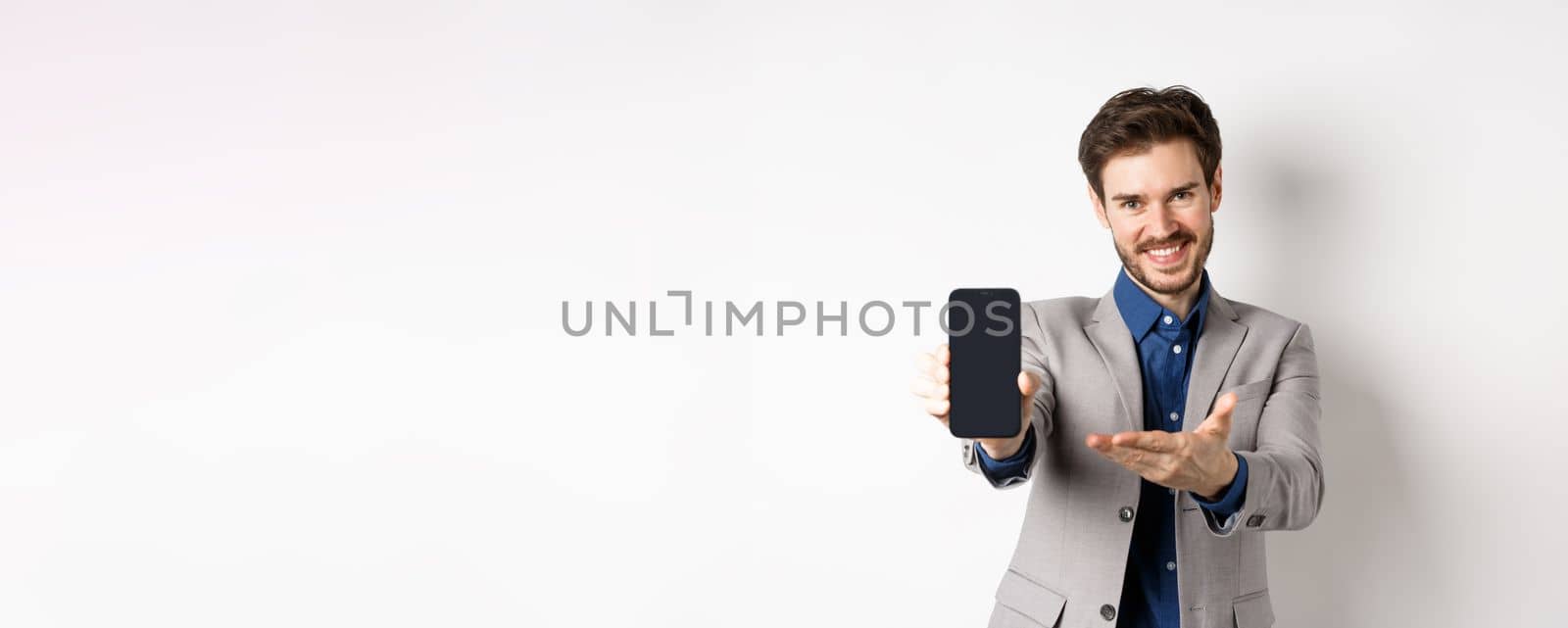 E-commerce and online shopping concept. Here you go. Smiling pleasant salesman demonstrate promo on phone, showing empty smartphone screen, white background.