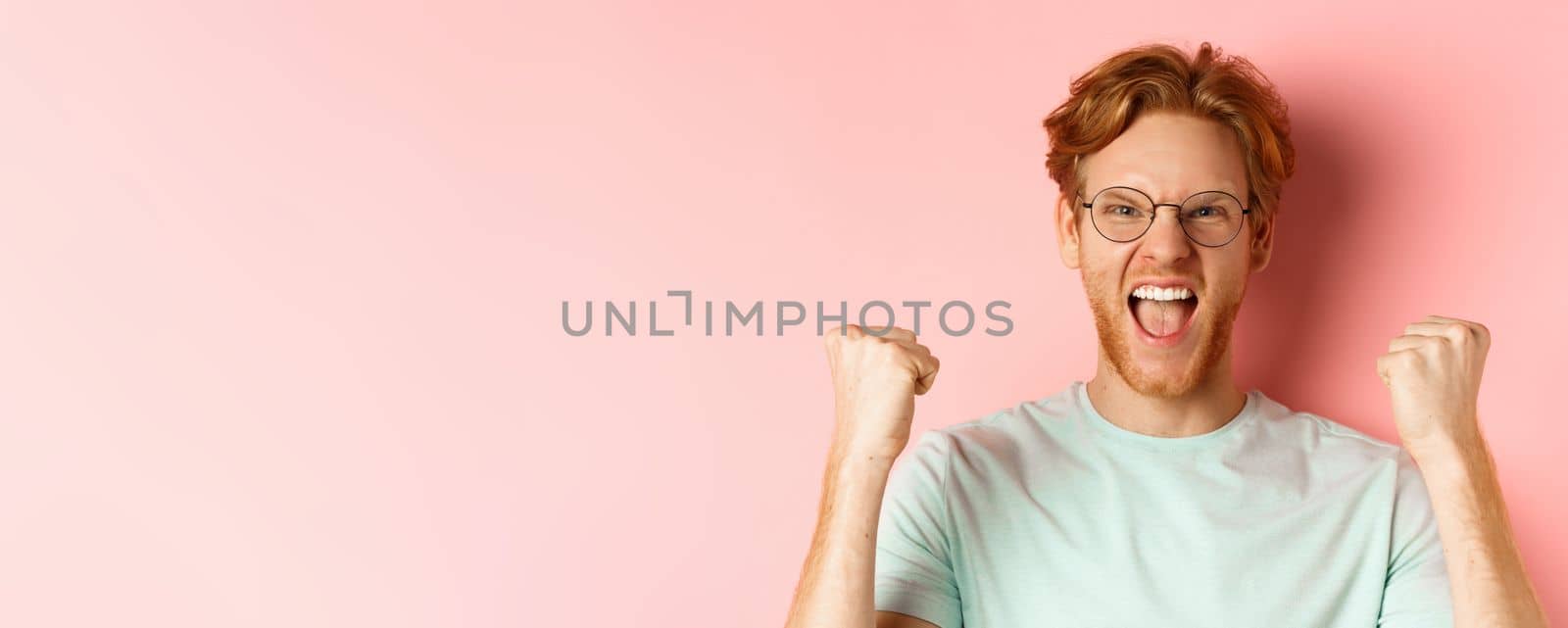 Close up of satisfied lucky redhead man winning, shouting from joy and making fist pump, celebratig victory, standing like champion over pink background.