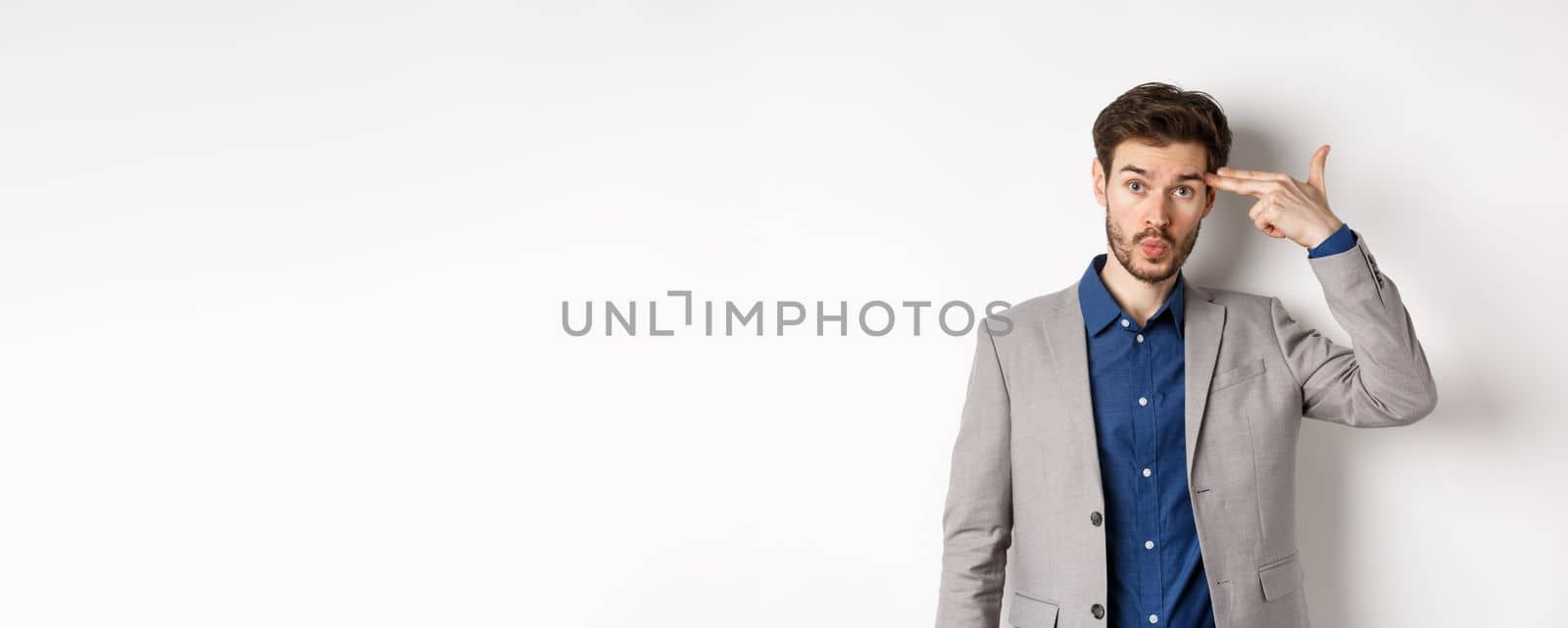 Annoyed guy in business suit shoot himself with hand gun near head, look distressed and tired after work, standing on white background.