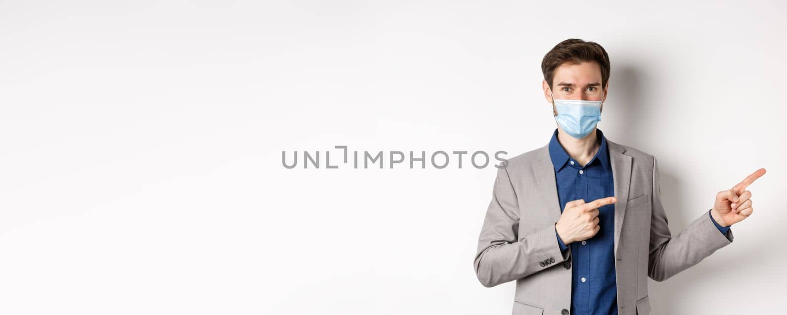 Covid-19, pandemic and business concept. Confident male manager in suit and medical mask showing way, pointing fingers right at advertisement, white background by Benzoix