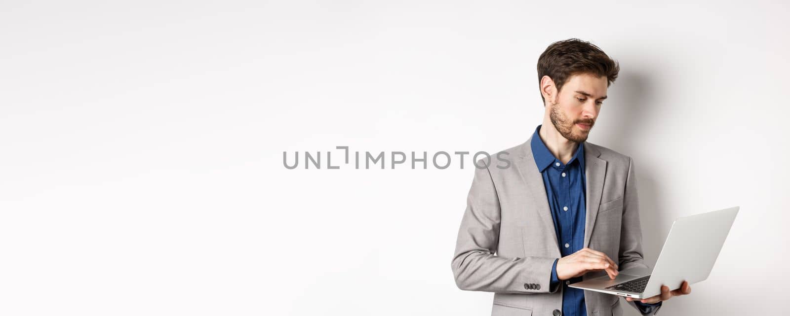 Handsome male entrepreneur working on laptop, looking serious at screen, standing against white background.