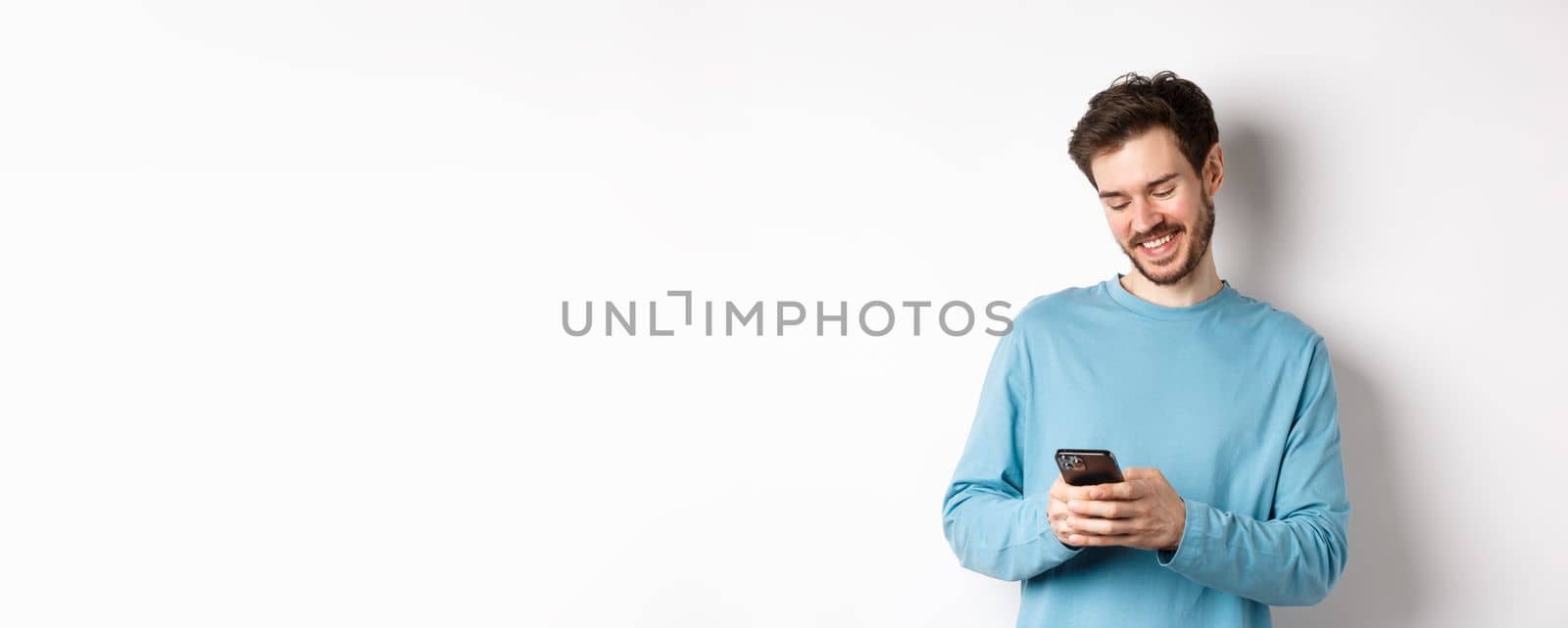 Portrait of modern caucasian man with beard, wearing casual clothes, reading smartphone screen and smiling, networking via mobile phone, standing over white background by Benzoix