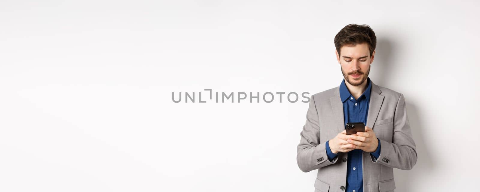 Businessman chatting on mobile phone, standing in suit with serious face, white background.