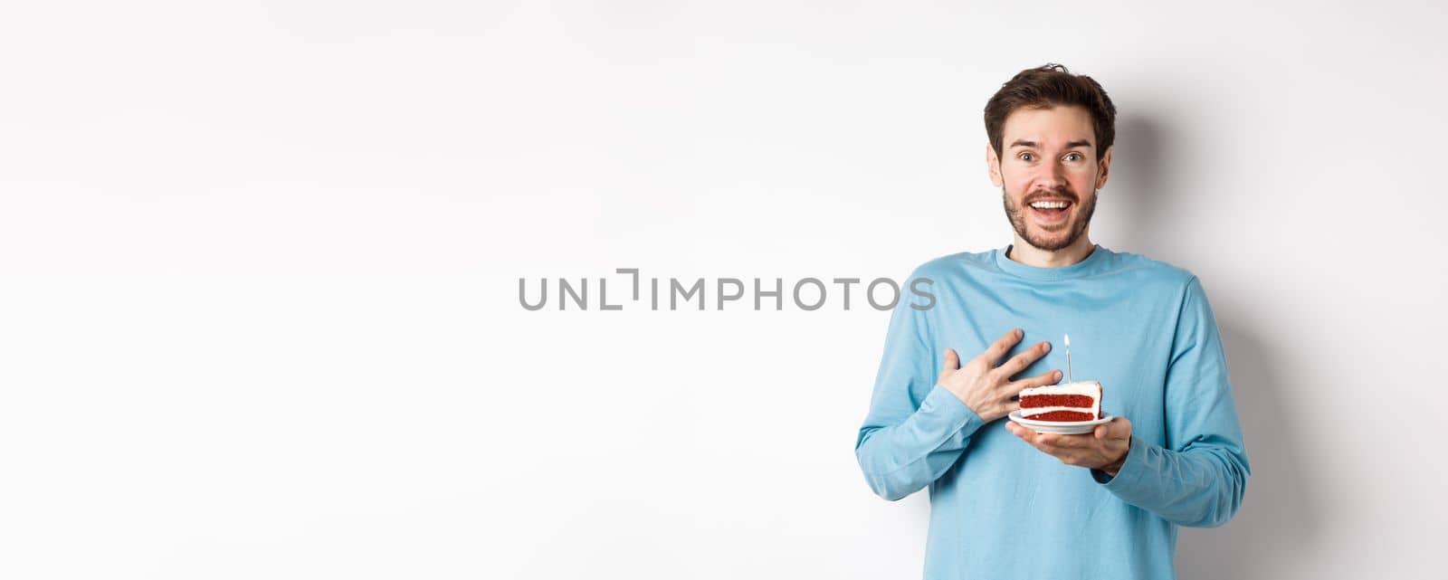 Surprised man receiving birthday cake with lit candle, being congratulated with b-day, gasping amazed, standing over white background by Benzoix