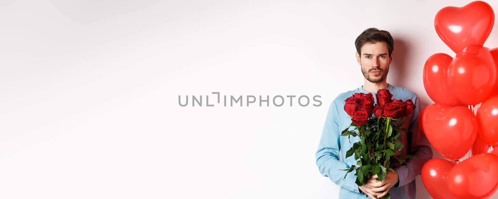 Valentines day romance. Confident young man holding bouquet of red roses, standing near hearts balloons, going on romantic date with lover, white background.