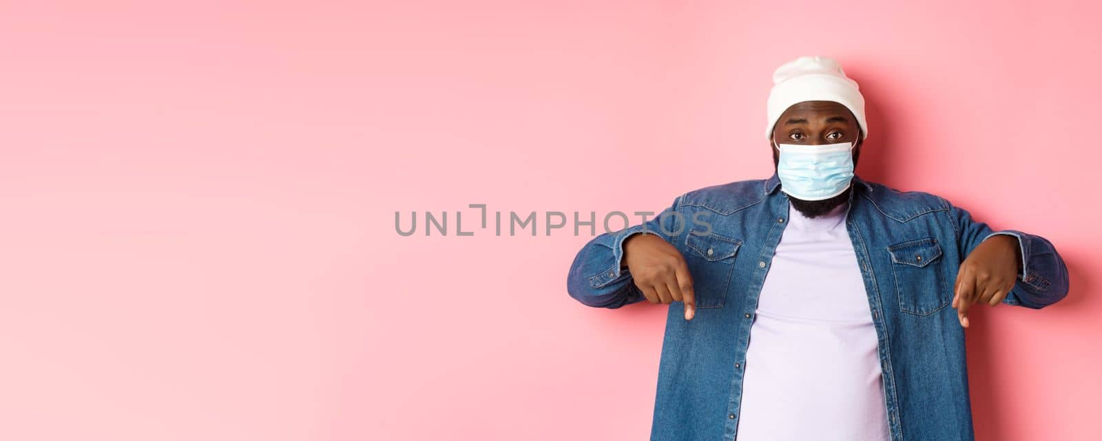 Covid-19, lifestyle and lockdown concept. Handsome african-american man in face mask, pointing fingers down, showing promo offer on bottom, standing over pink background.