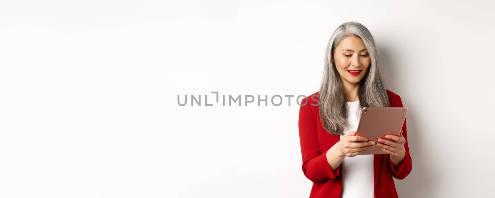 Business. Successful senior businesswoman working with digital tablet, reading screen and smiling, standing in elegant red blazer over white background by Benzoix