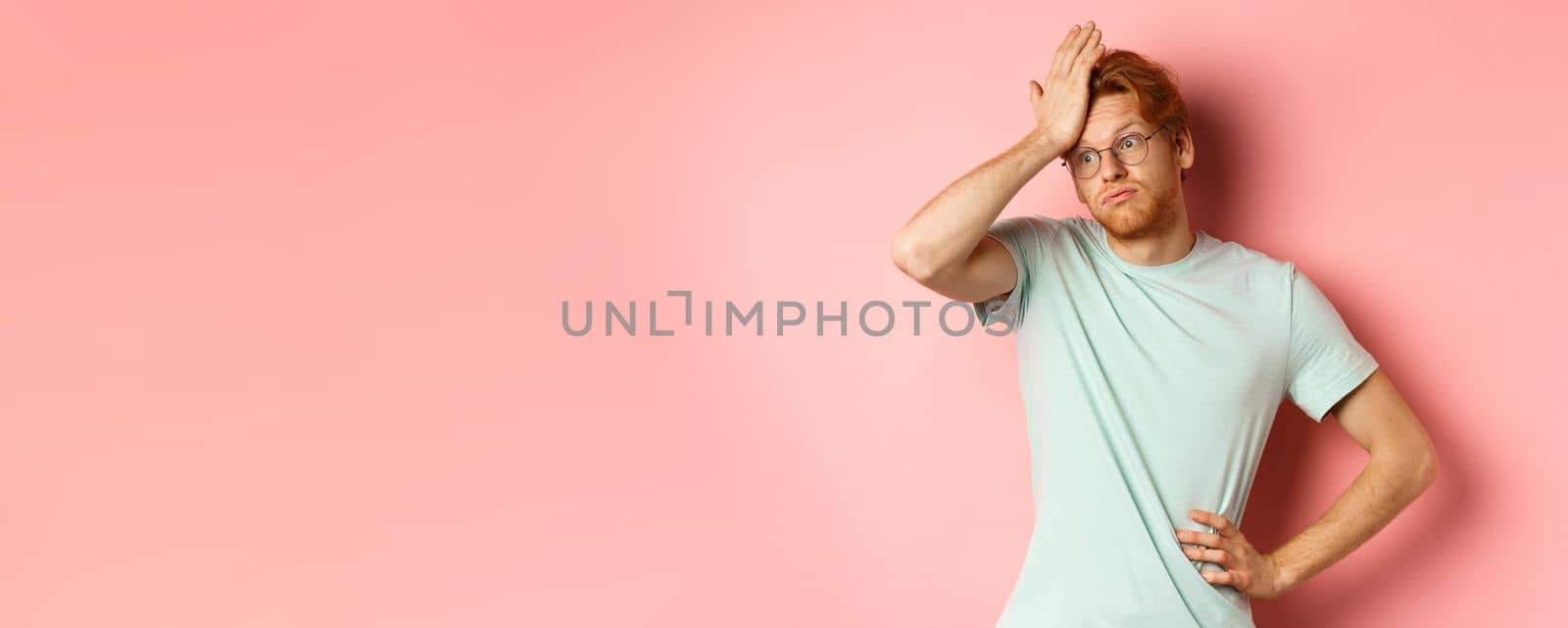 Tired young man with red hair, wearing glasses, looking annoyed and tensed, making facepalm gesture and exhale bothered, standing over pink background.