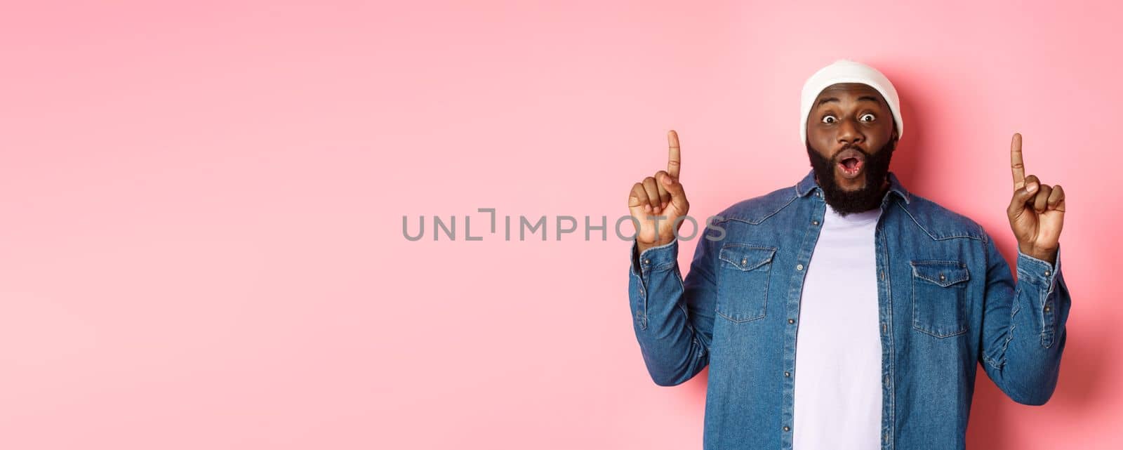 Excited and impressed Black man showing amazing promo, pointing fingers up at logo, standing against pink background by Benzoix