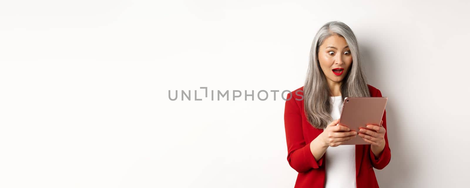 Business. Asian senior woman staring at digital tablet screen with amazed and surprised face, reading amazing news online, standing over white background.
