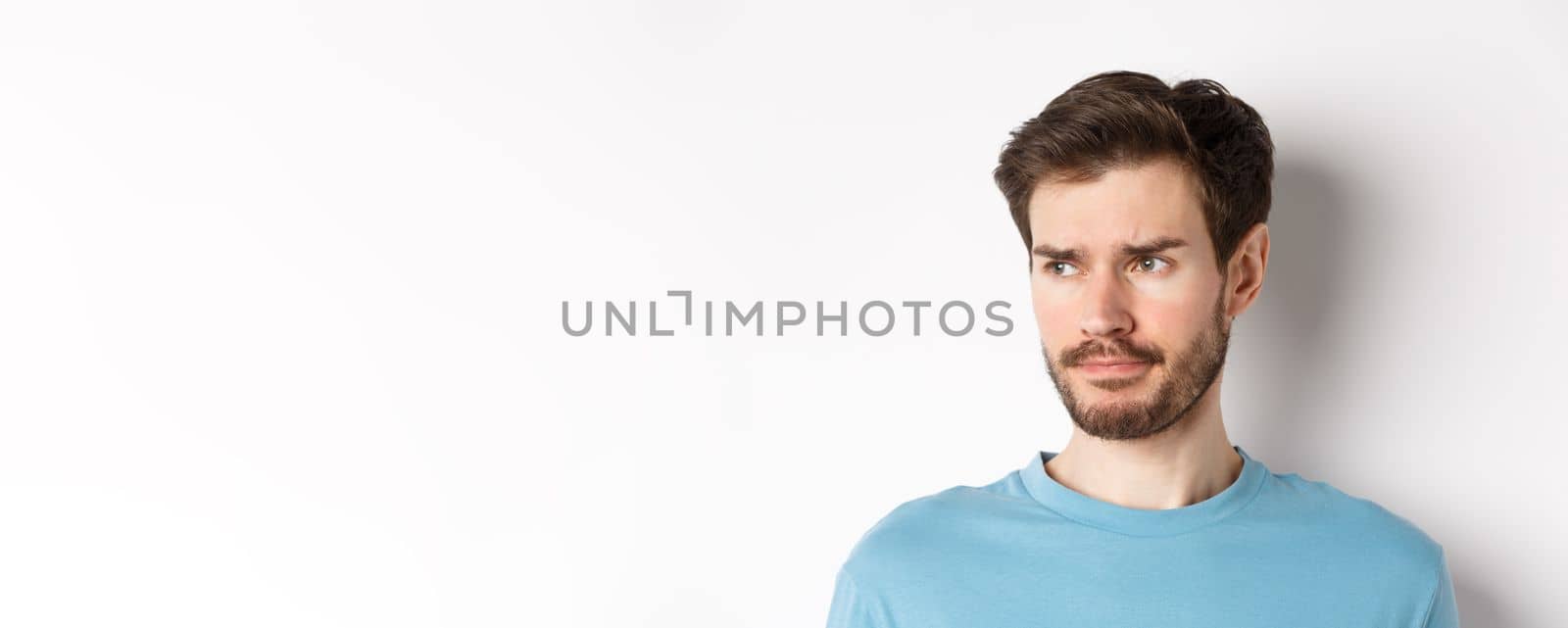 Close-up of disappointed sad man, grimacing and frowning, looking left upset, standing doubtful against white background by Benzoix