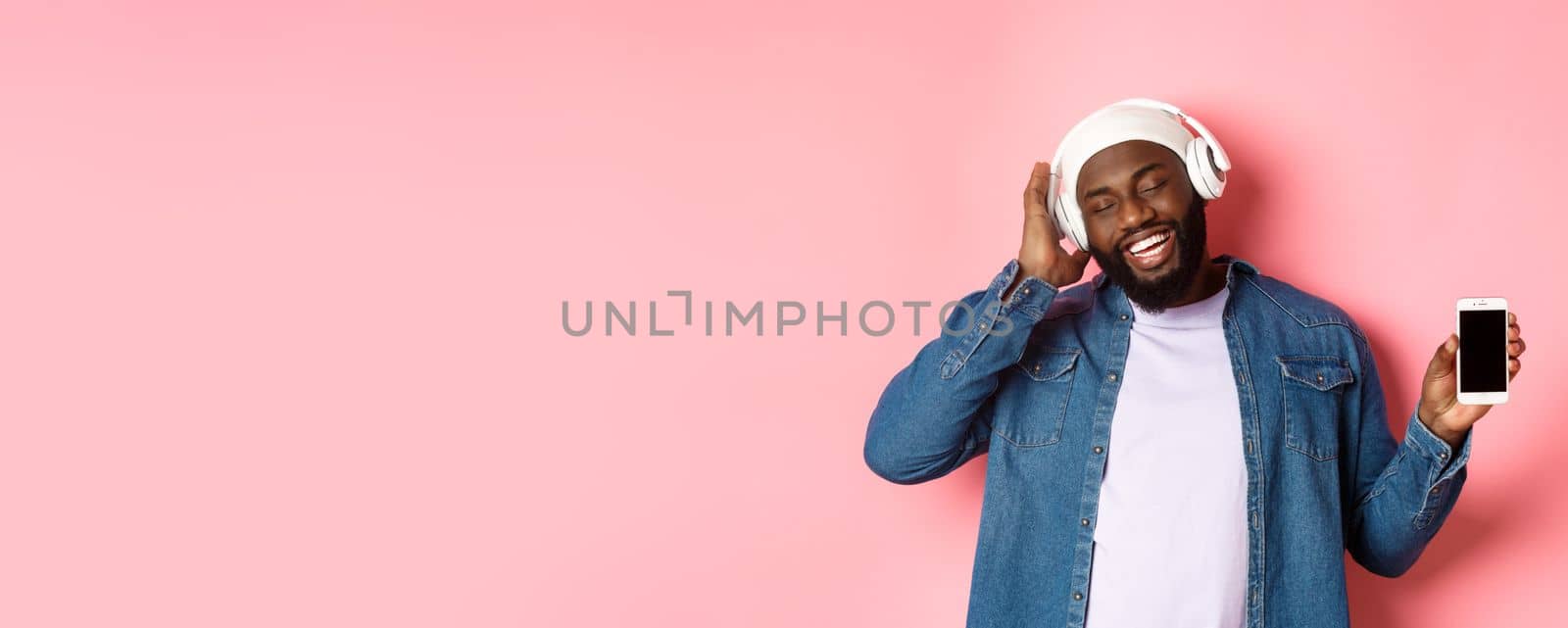 Carefree handsome Black man listening music in headphones, enjoying sound and showing smartphone screen, standing with pleased smile on face against pink background by Benzoix