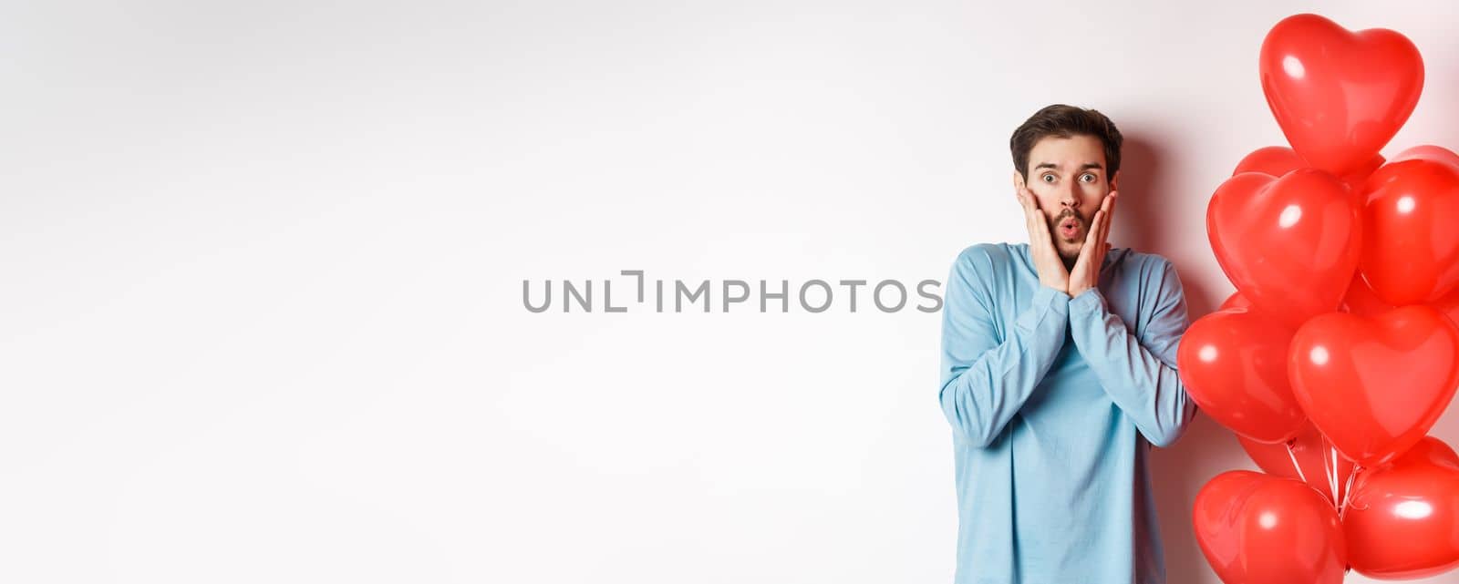 Valentines day. Image of young man standing near hearts balloons with shocked face, staring startled at camera, white background by Benzoix