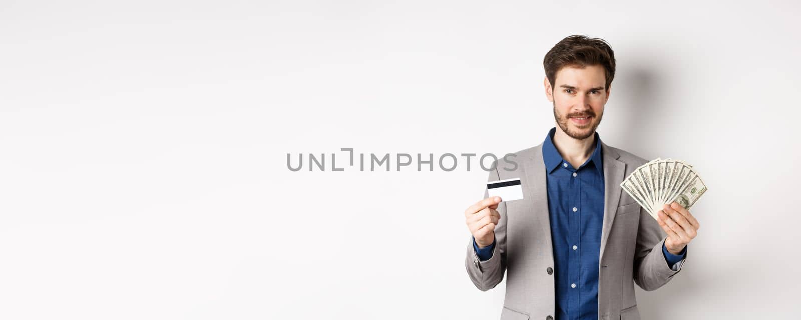 Rich smiling man in suit showing money and plastic credit card, standing with dollar bills and look pleased, white background by Benzoix