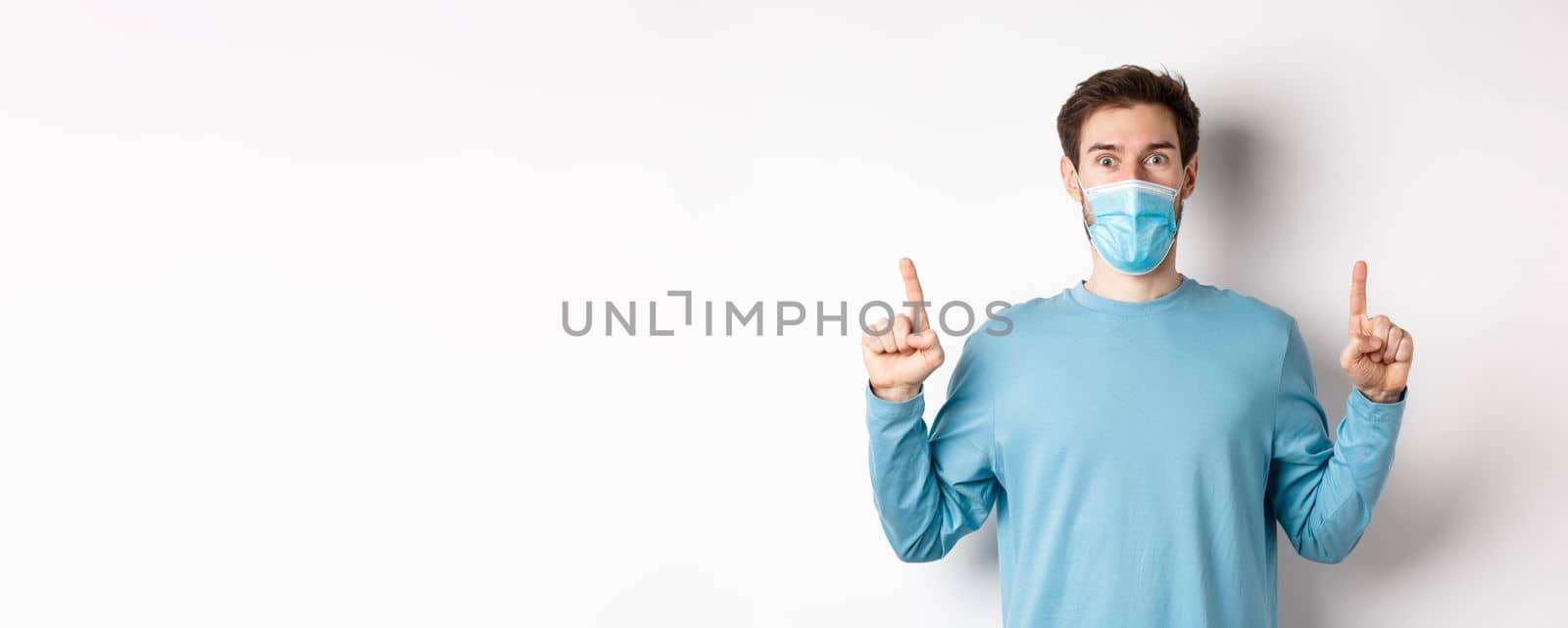 Coronavirus, health and quarantine concept. Excited smiling guy checking out special deal, pointing fingers up, stare amazed at camera, wear medical mask on white background.