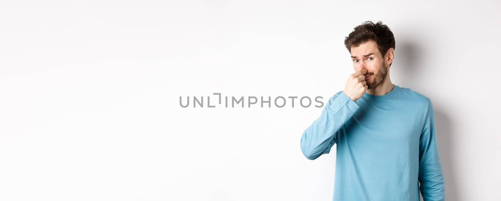 Disgusted young man in sweatshirt shut his nose, looking at something with bad smell, standing over white background.