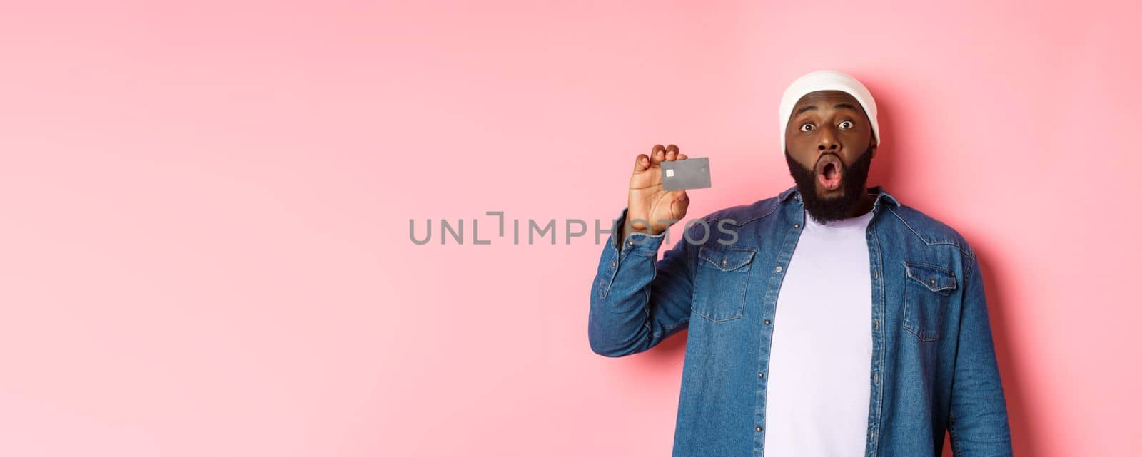 Shopping concept. Amazed Black guy showing credit card, staring at camera impressed, recommending bank, standing over pink background by Benzoix