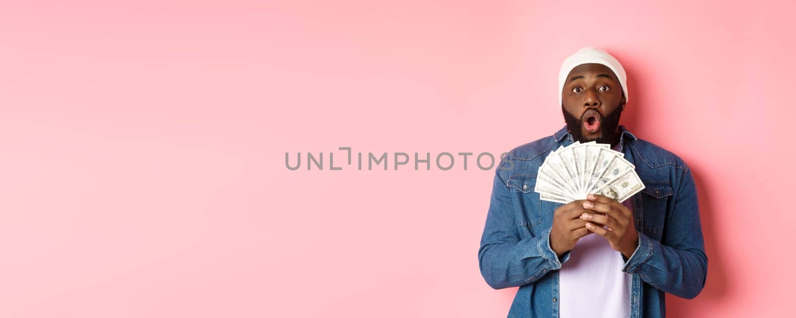 Amazed african-american man receive cash prize, showing money and staring at camera in awe, standing over pink background by Benzoix