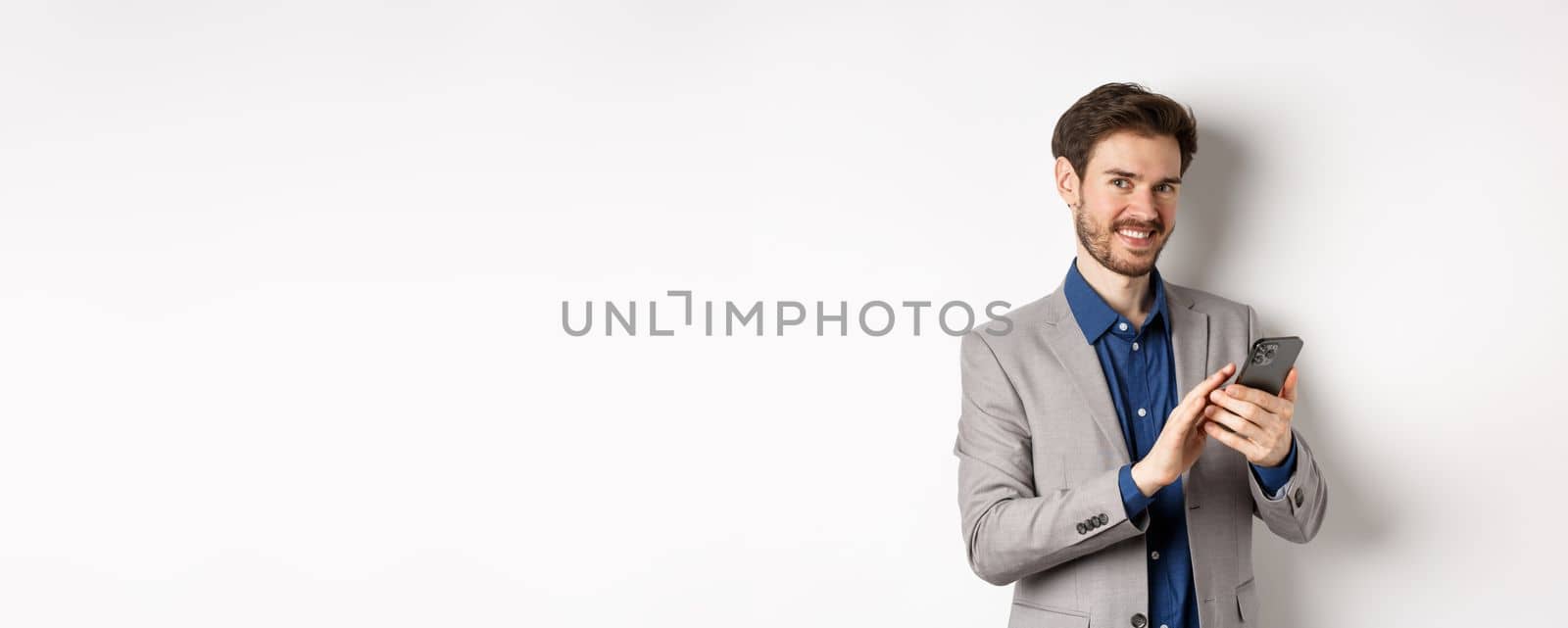 Smiling successful business man using smartphone on white background, standing in suit.