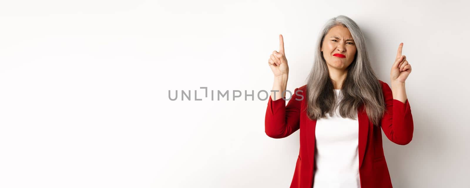 Skeptical and disappointed asian businesswoman pointing fingers up, grimacing and showing bad promo offer, standing over white background.