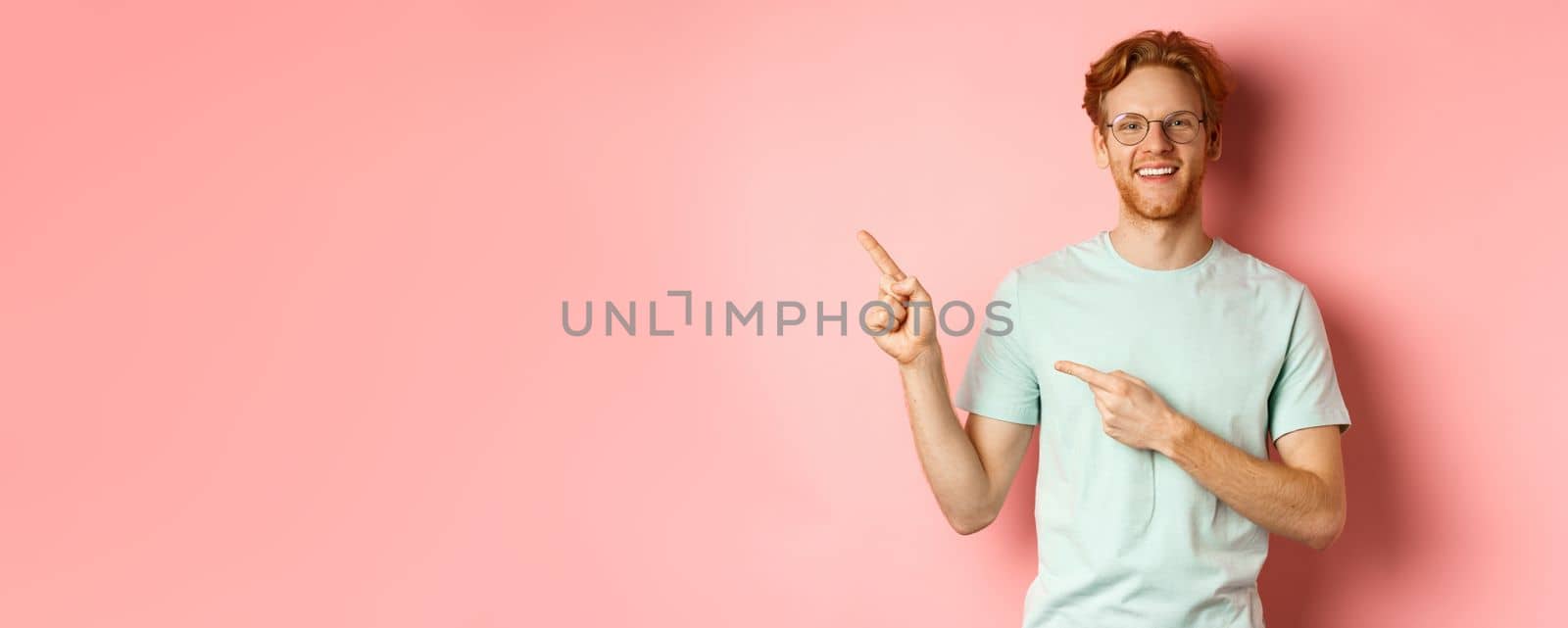 Portrait of cheerful young man with red hair, wearing glasses, pointing fingers at upper left corner and smiling, standing over pink background by Benzoix