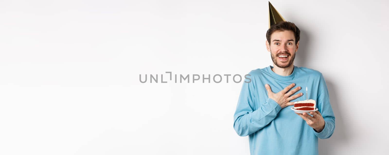 Celebration and holidays concept. Surprised birthday boy in party hat, holding bday cake and looking thankful, standing over white background by Benzoix
