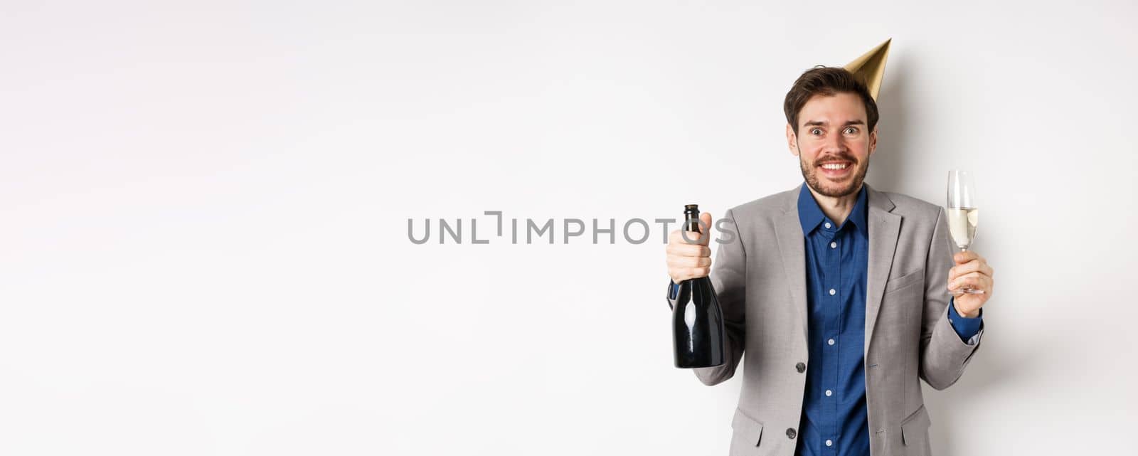 Celebration and holidays concept. Excited handsome birthday guy in party hat smiling, drinking champagne and having fun, white background.