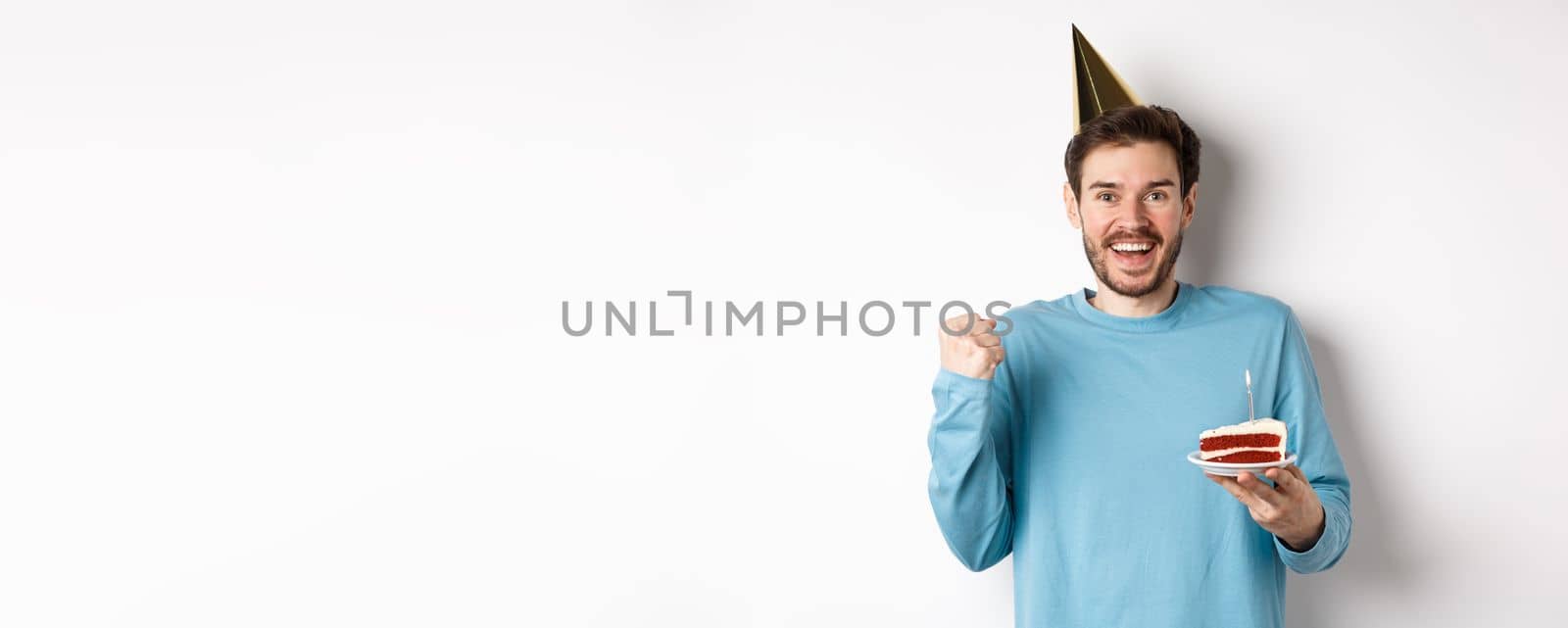 Celebration and holidays concept. Cheerful young man celebrating birthday in party hat, saying yes and fist pump in joy, holding bday cake, white background.