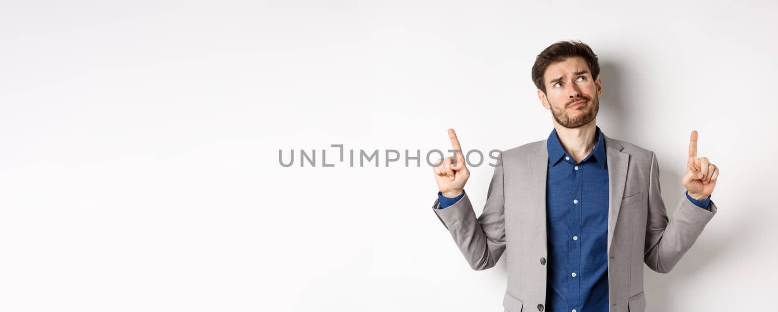 Skeptical and hesitant young businessman in suit looking up with disappointed smirk, feel unsure about deal, standing dissatisfied on white background.