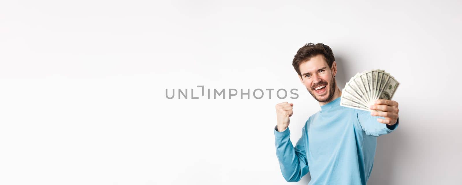 Happy caucasian man stretch out hand with money in dollars, saying yes and celebrating income, got cash prize, standing over white background.