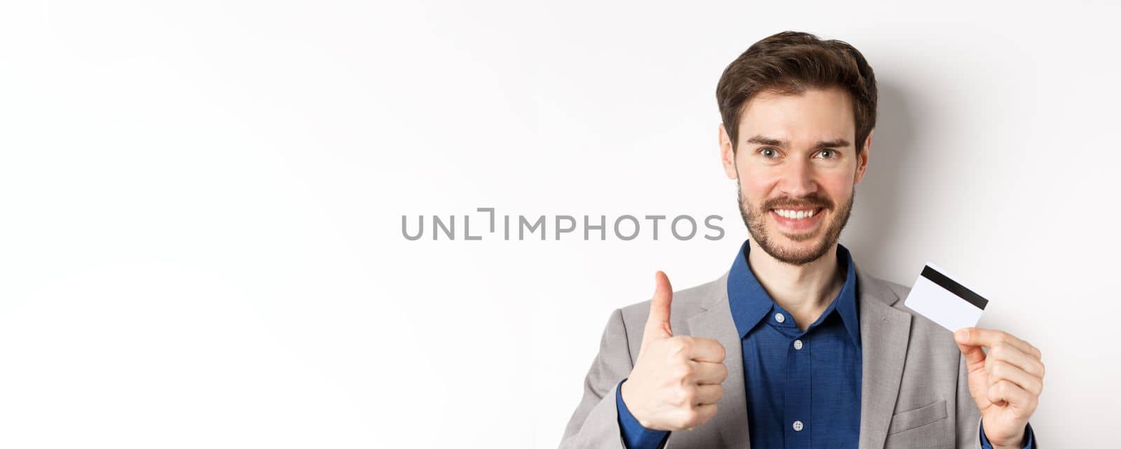 Shopping. Handsome satisfied client showing thumbs up and plastic credit card, smiling pleased, white background by Benzoix