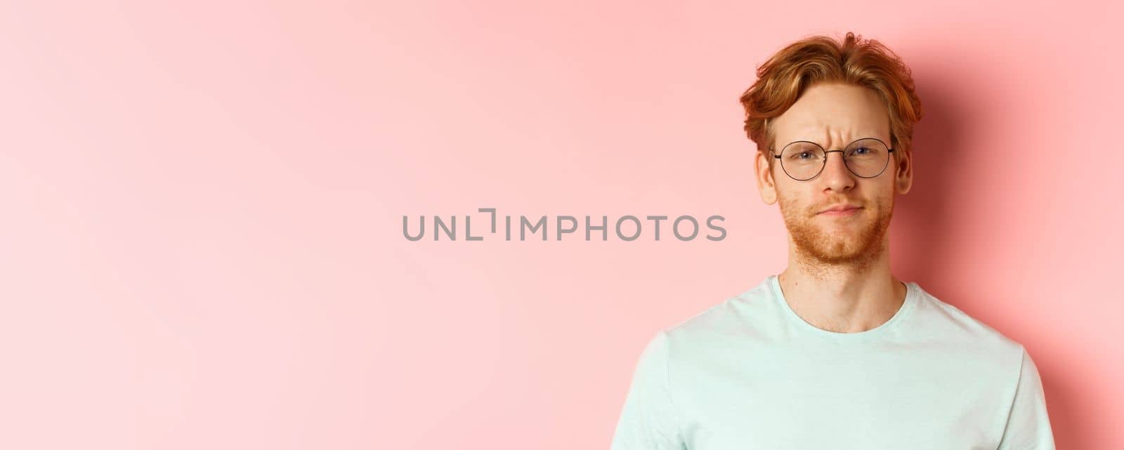 Headshot of skeptical redhead man in glasses and t-shirt frowning disappointed, staring with disapproval and judgement, standing over pink background.