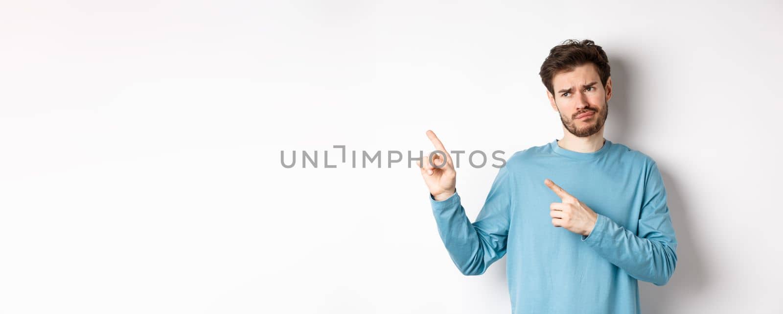 Skeptical and unimpressed young man grimacing, pointing fingers and looking at bad promotion, standing dissatisfied over white background.