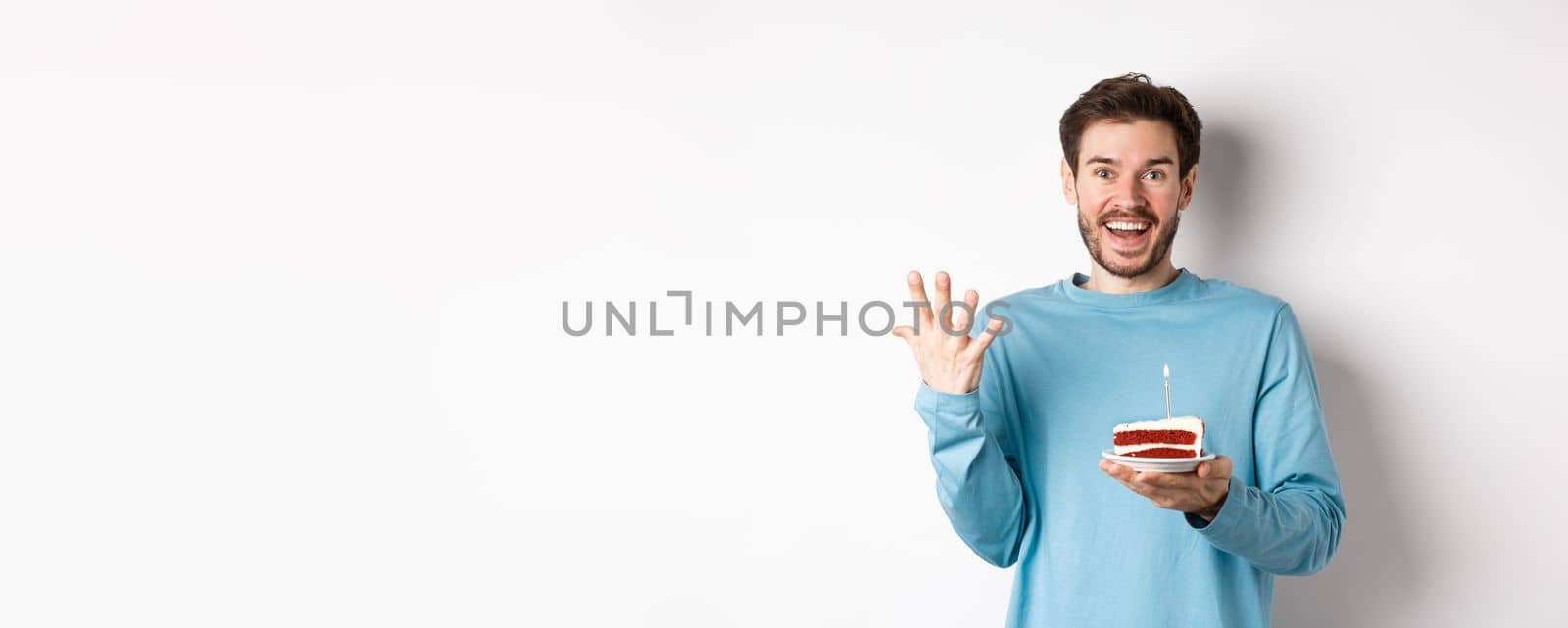 Excited man receive birthday surprise, holding bday cake and smiling happy, standing over white background, making wish on lit candle.