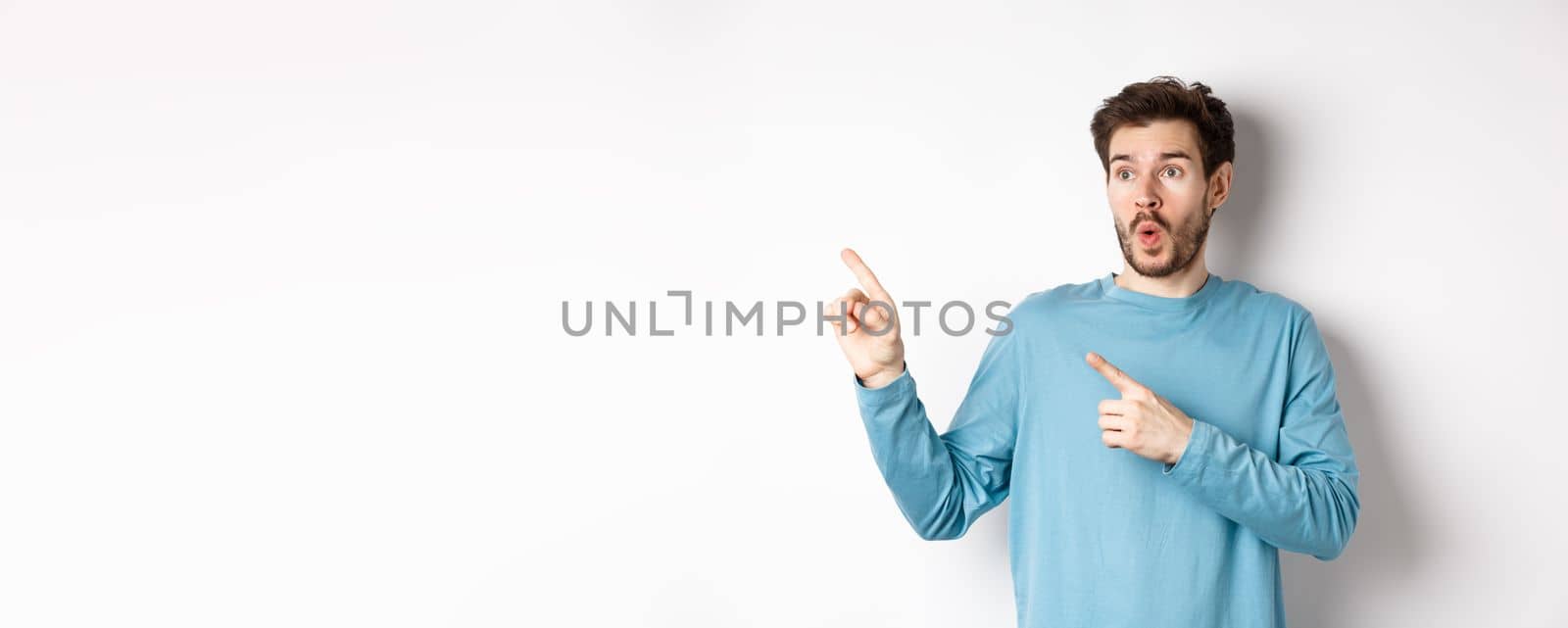 Wow check it out. Impressed young man pointing and looking left at special promo offer or discounts on white background, gasping amazed, standing in studio.