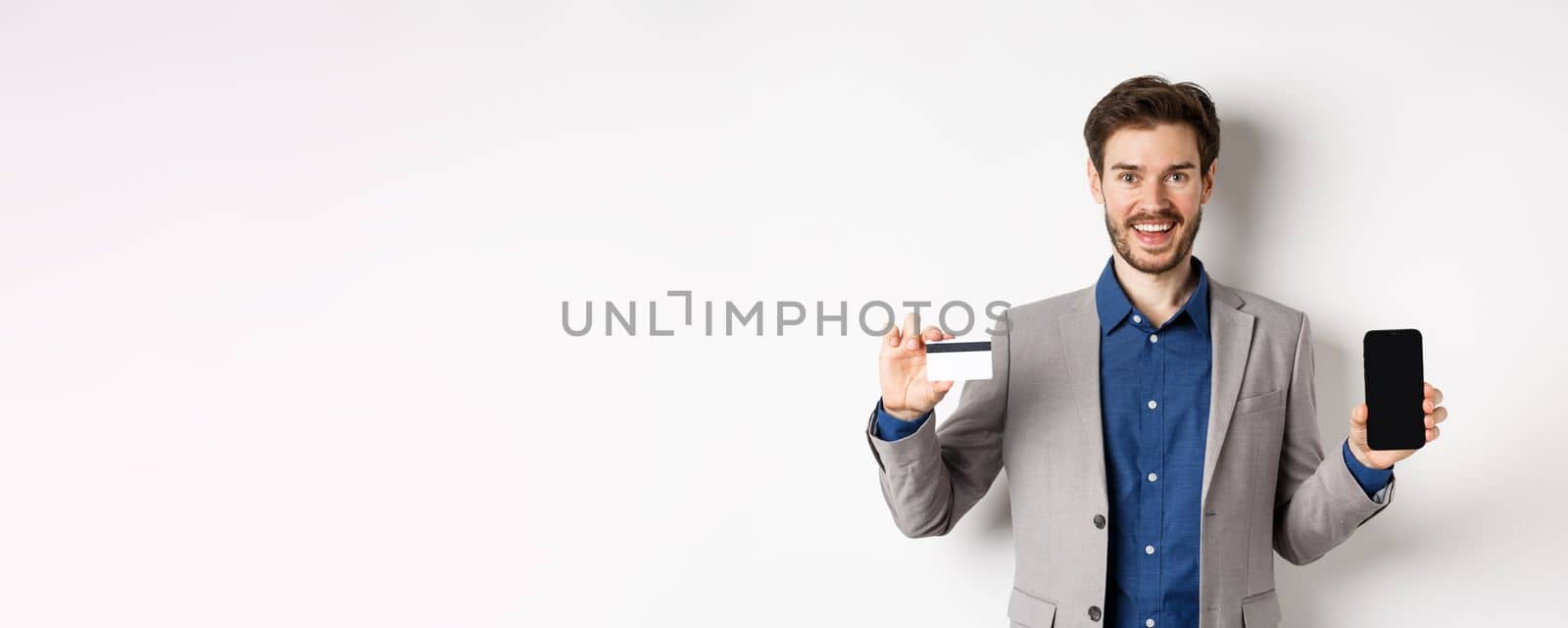 Online shopping. Smiling business man in suit showing plastic credit card with empty smartphone screen, standing against white background.