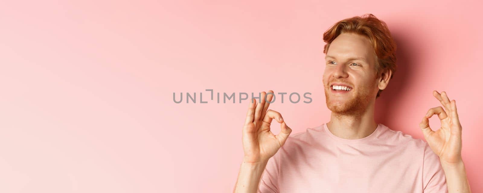 Close up of handsome redhead man in t-shirt, smiling satisfied and looking left, showing okay signs in approval, praising awesome product, standing over pink background by Benzoix