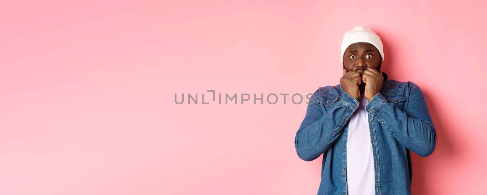 Scared Black guy trembling from fear, biting fingernails and staring at camera, standing over pink background.