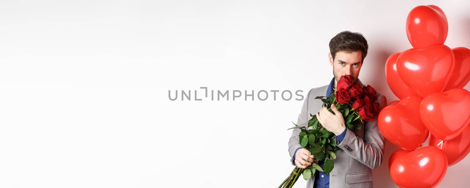 Romantic man smell bouquet of red roses and looking passionate at camera. Boyfriend in suit going on Valentines date with gifts and heart balloons, white background.