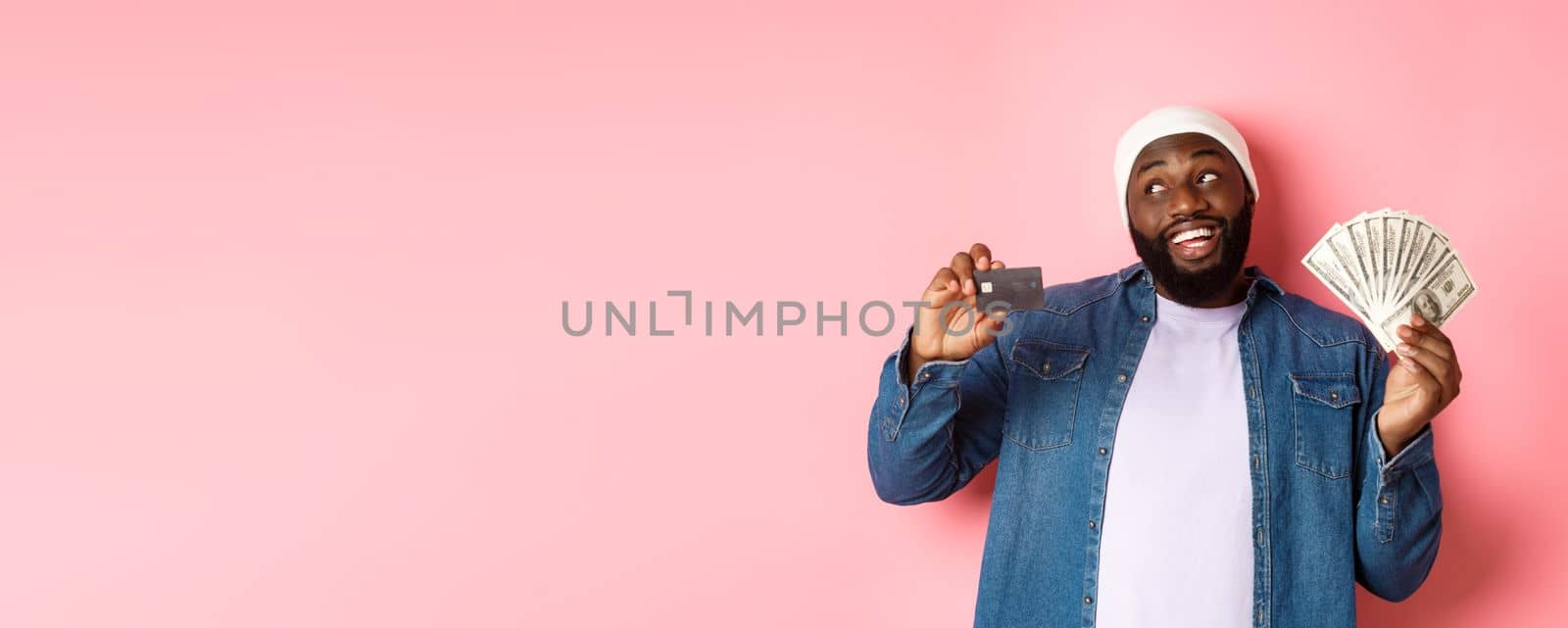 Dreamy african-american man showing credit card and dollars, thinking about shopping and smiling, standing over pink background.