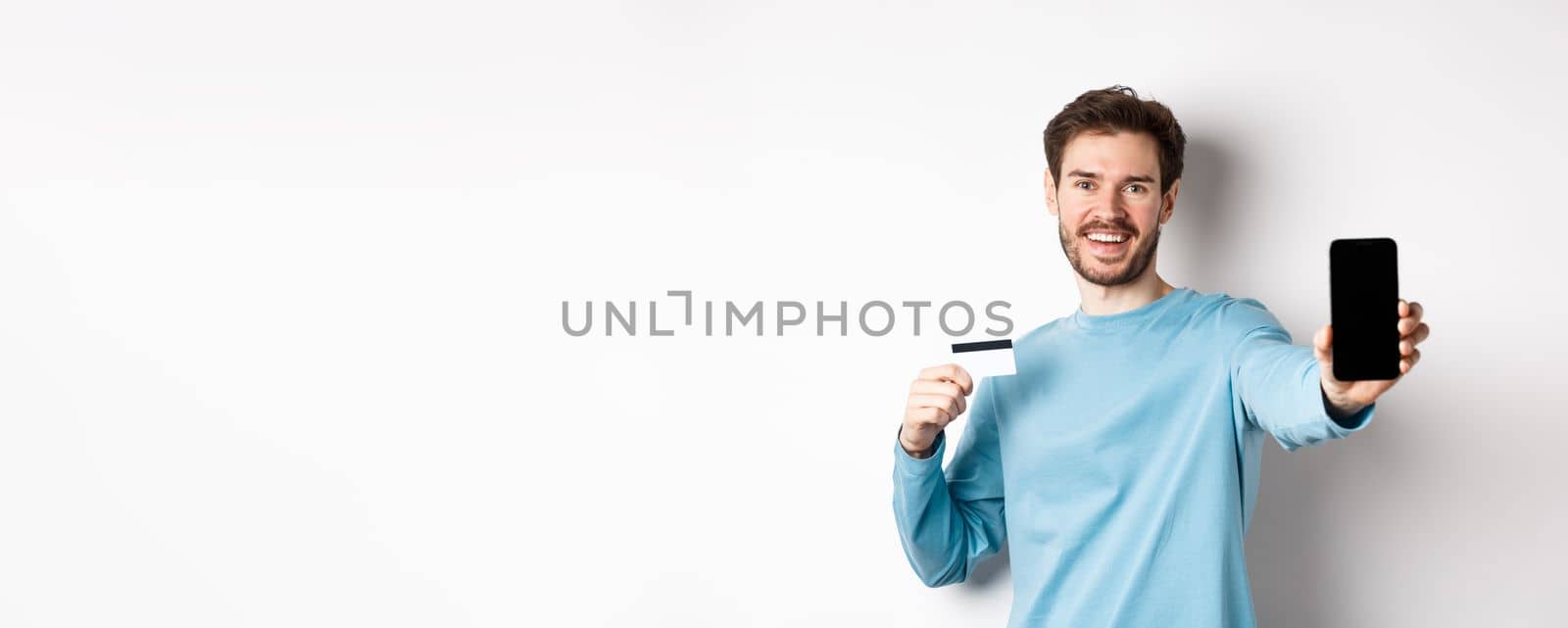 E-commerce and shopping concept. Smiling caucasian man showing plastic credit card and empty smartphone screen, recommending online app, white background by Benzoix
