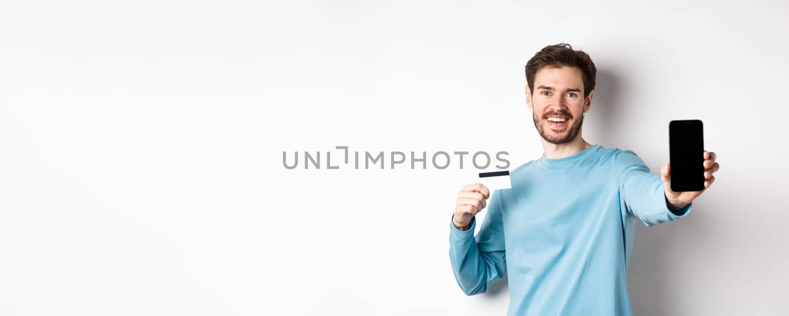 E-commerce and shopping concept. Smiling caucasian man showing plastic credit card and empty smartphone screen, recommending online app, white background.