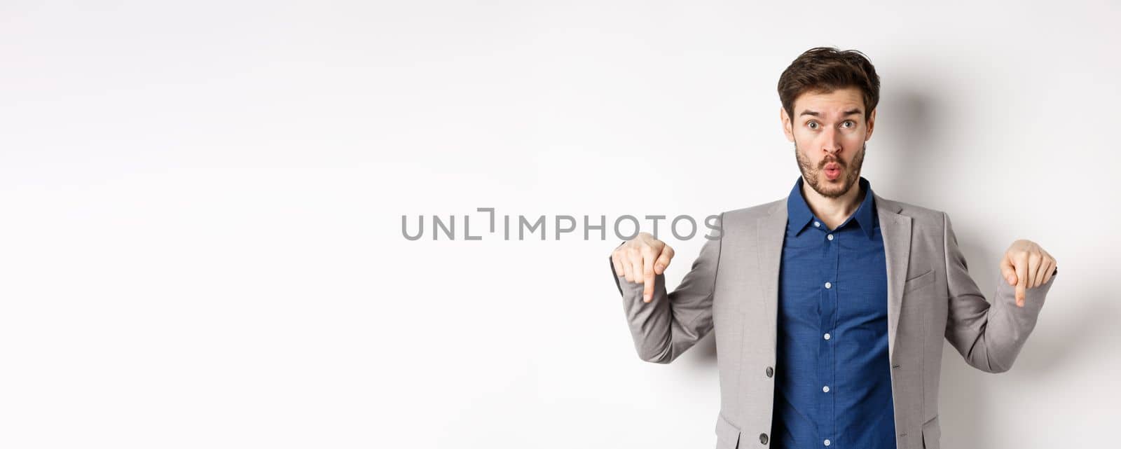Wow look down. Excited business guy in suit pointing fingers bottom advertisement and look amazed, standing on white background.