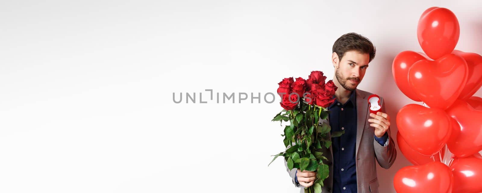 Handsome boyfriend in suit making proposal on lovers day, holding engagement ring and red roses, prepare surprise flowers and heart balloons for girlfriend on Valentines, white background.