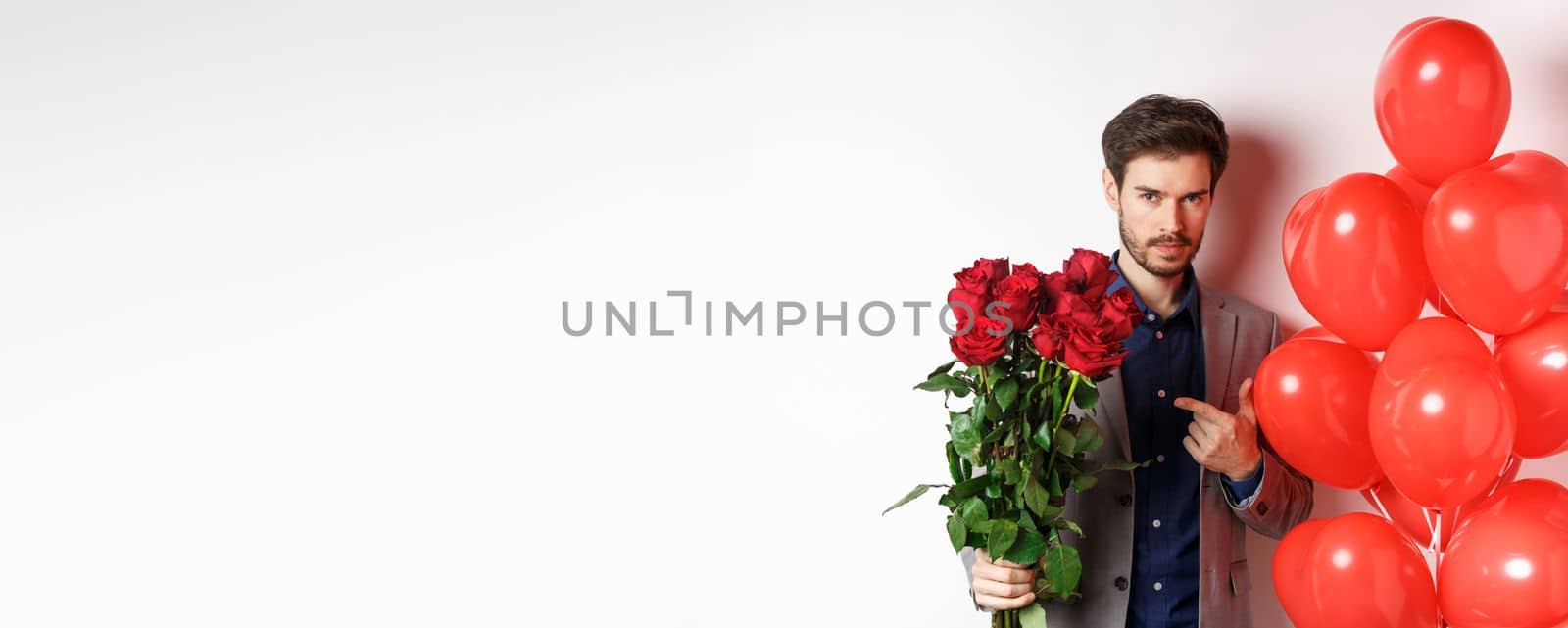 Handsome man in suit going on romantic Valentines day date, pointing at bouquet of roses, standing near heart balloon over white background by Benzoix