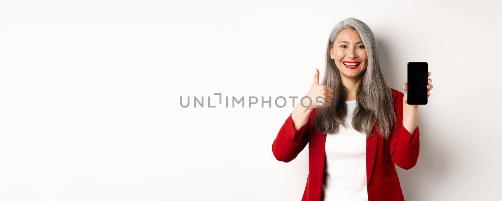 Satisfied asian elderly businesswoman showing blank smartphone screen and thumb-up, praising online promotion or company app, standing over white background.