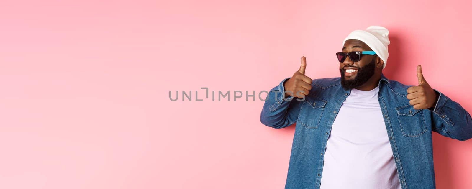 Happy and satisfied african-american guy in sunglasses showing thumbs-up, praise and like promo, agree with you, standing over pink background by Benzoix