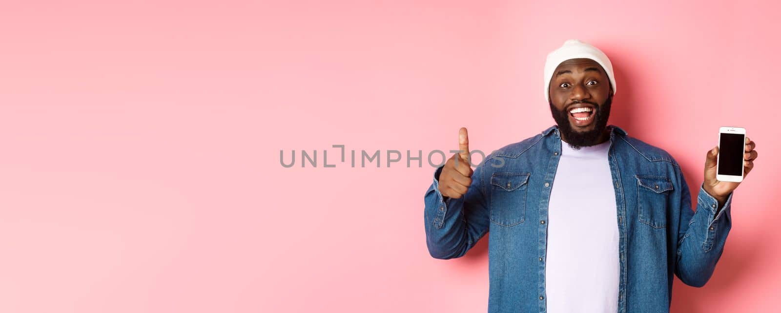 Online shopping and technology concept. Happy african-american hipster man showing thumbs-up and mobile screen, praising app, standing over pink background.